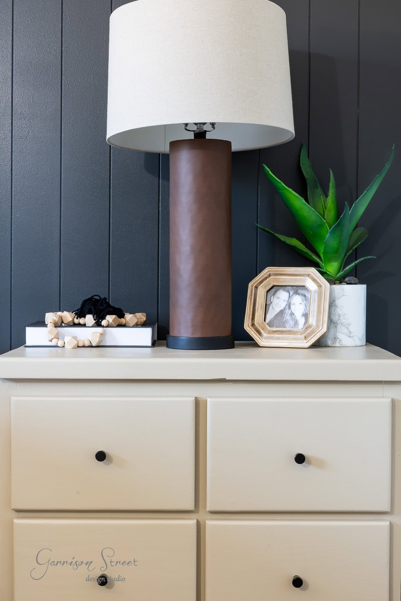 A beige dresser with black knobs stands against a dark wall. On top, there's a brown cylindrical lamp with a white shade, framed photos, a green plant, and decorative items like a bracelet and a small book.