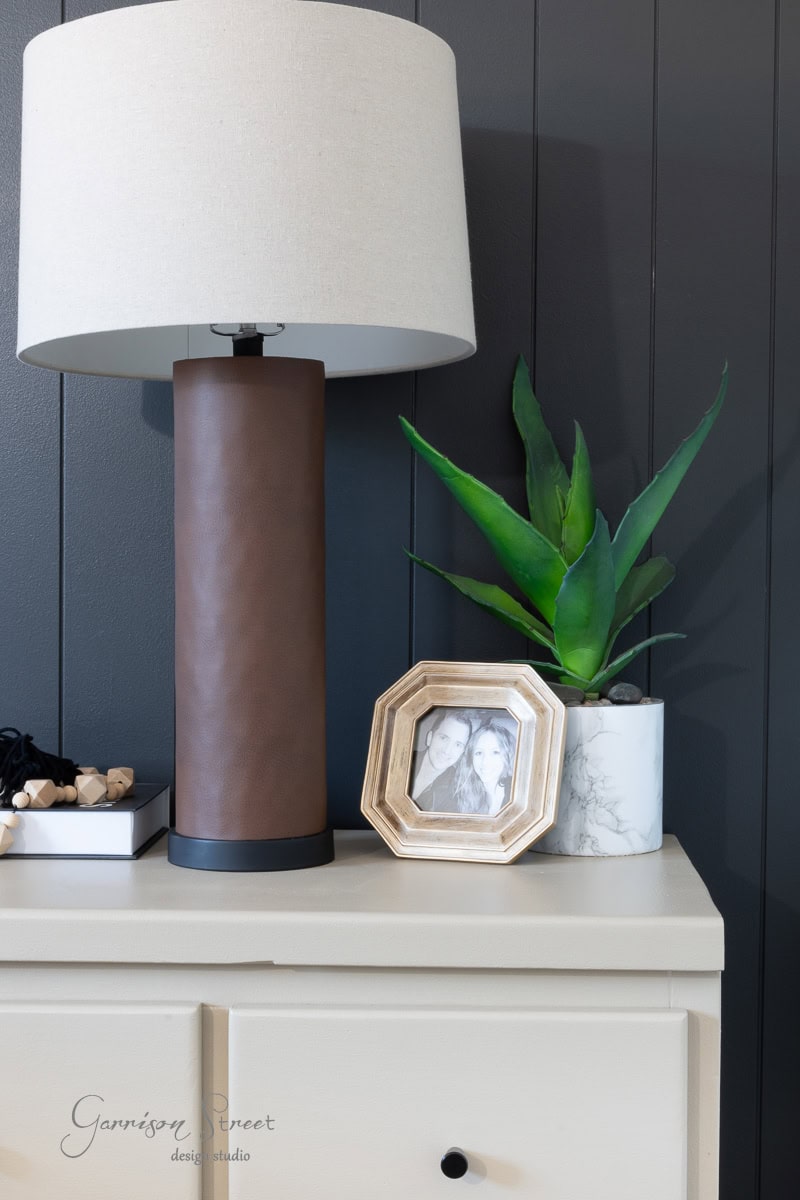 A modern table lamp with a beige shade sits on a light-colored dresser. Next to it are a small potted plant and a framed black-and-white photo. A dark paneled wall serves as the background.