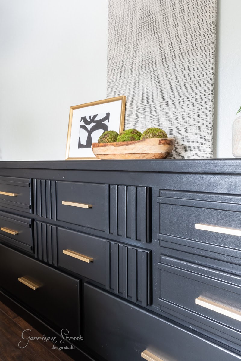 A stylish black dresser with gold handles is adorned with a wooden bowl of green decorative orbs and a framed abstract art piece. A textured panel hangs on the wall above, complementing the modern decor. The dresser is labeled "Garrison Street design studio."
