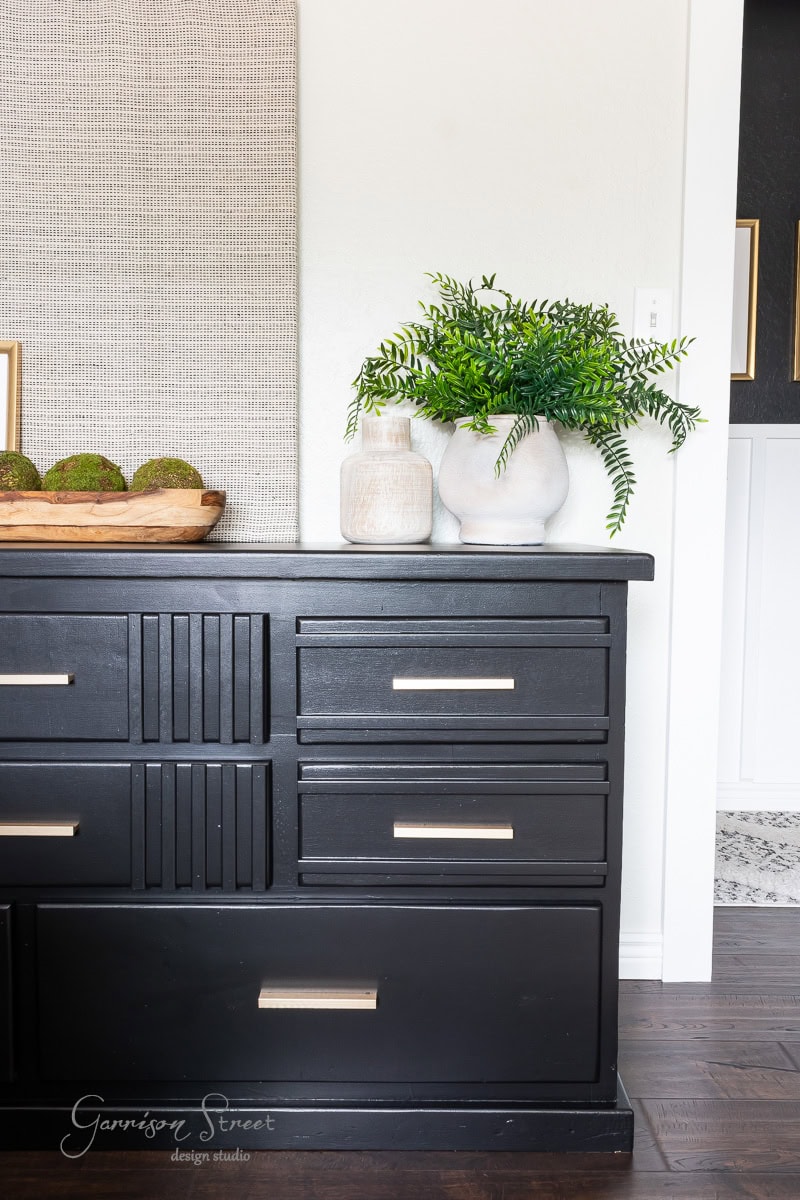 A black dresser with silver handles is adorned with a wooden tray of moss balls and two white vases filled with green foliage. The backdrop features a light-colored wall, adding contrast to the dark furniture.