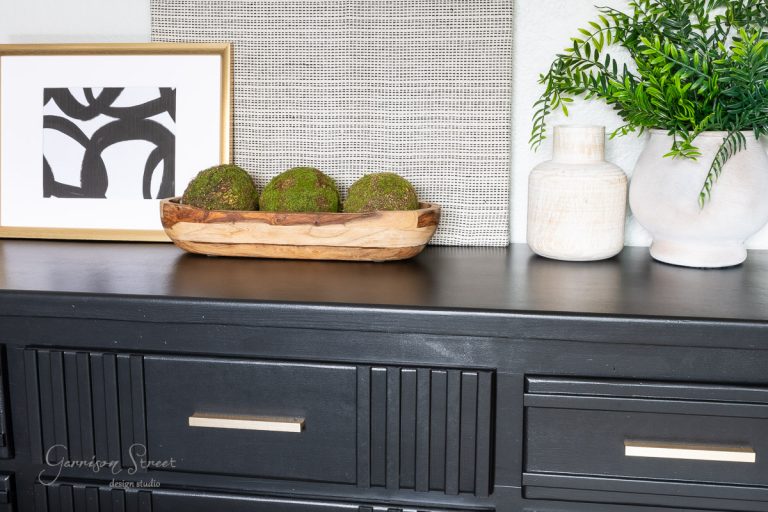 A black dresser from a DIY makeover showcases a modern art piece, a wooden bowl with moss spheres, and decorative vases with greenery. A textured square panel completes the arrangement, offering a minimalist and contemporary aesthetic.