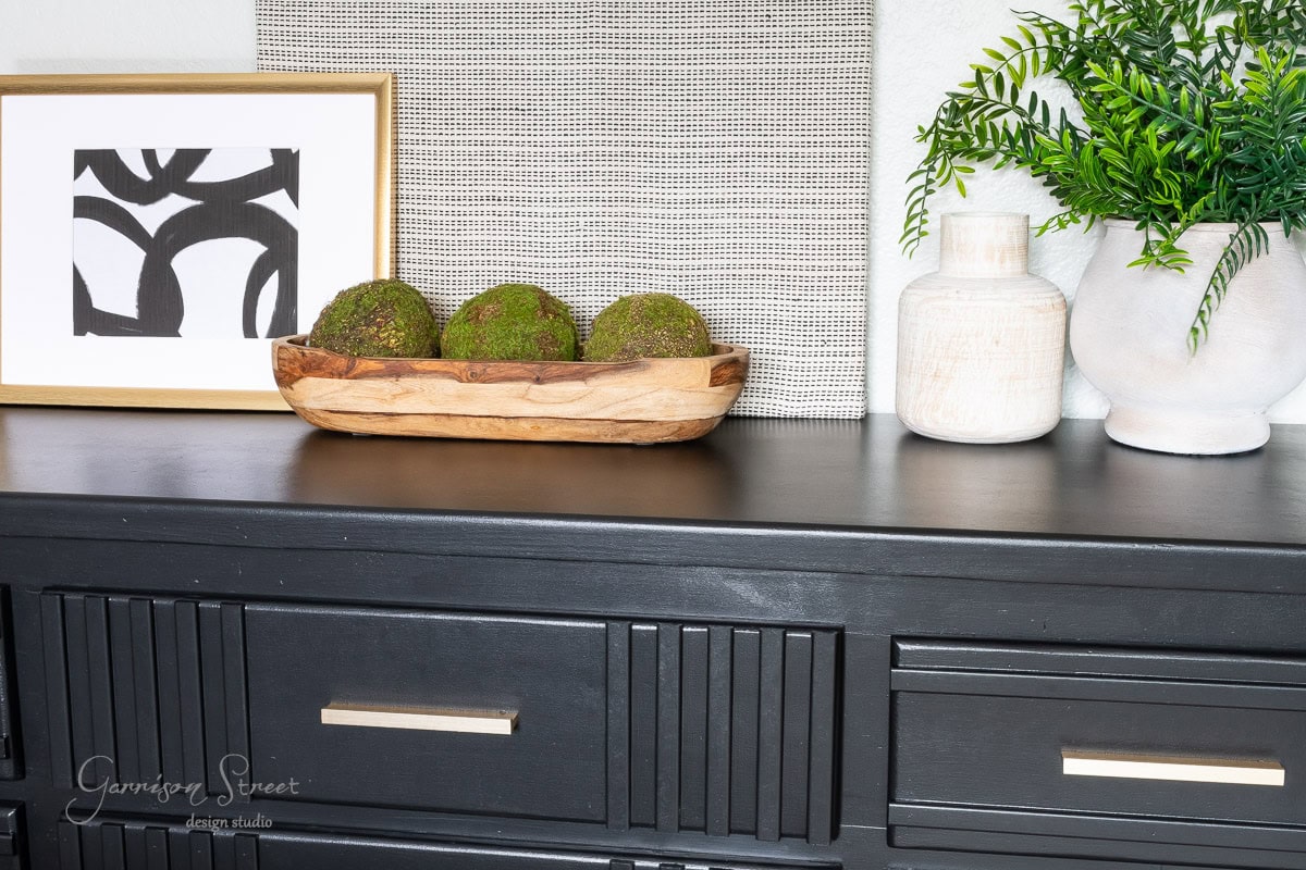 A black dresser from a DIY makeover showcases a modern art piece, a wooden bowl with moss spheres, and decorative vases with greenery. A textured square panel completes the arrangement, offering a minimalist and contemporary aesthetic.