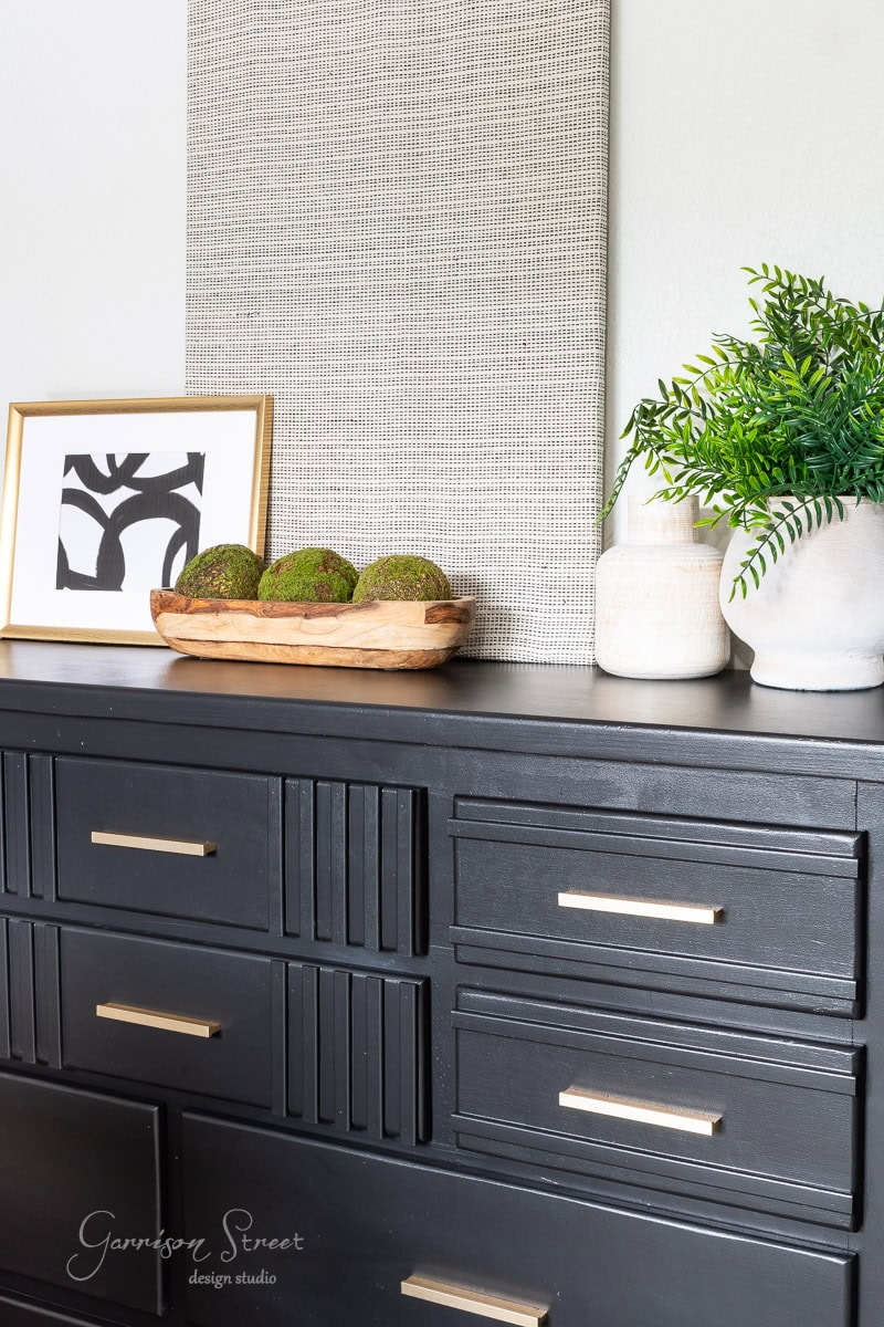 A black dresser features gold handles and displays a wooden tray with green moss balls, white vases with plants, and a framed abstract art piece. In the background, a textured wall panel adds a modern touch.