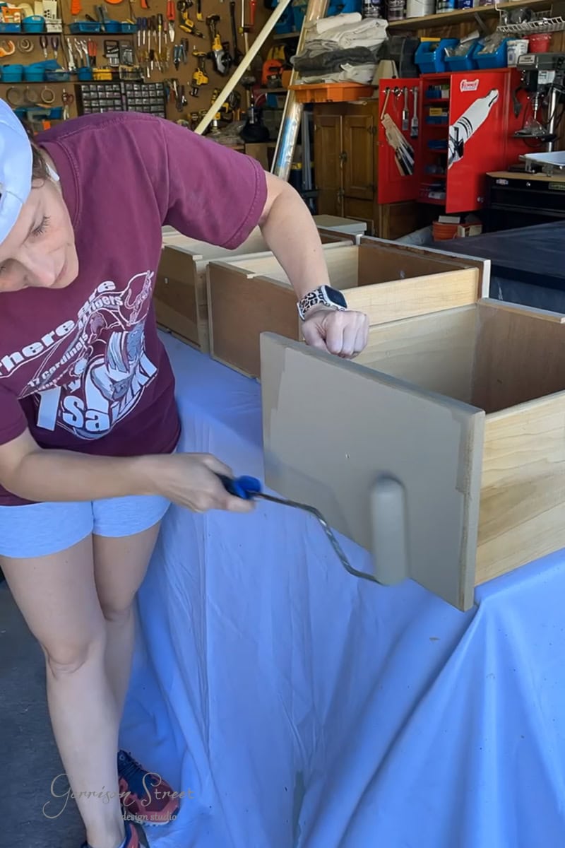 A person in a maroon t-shirt and white shorts uses a gray paint roller on a wooden drawer front. They're working on a covered surface in a workshop filled with tools and equipment.