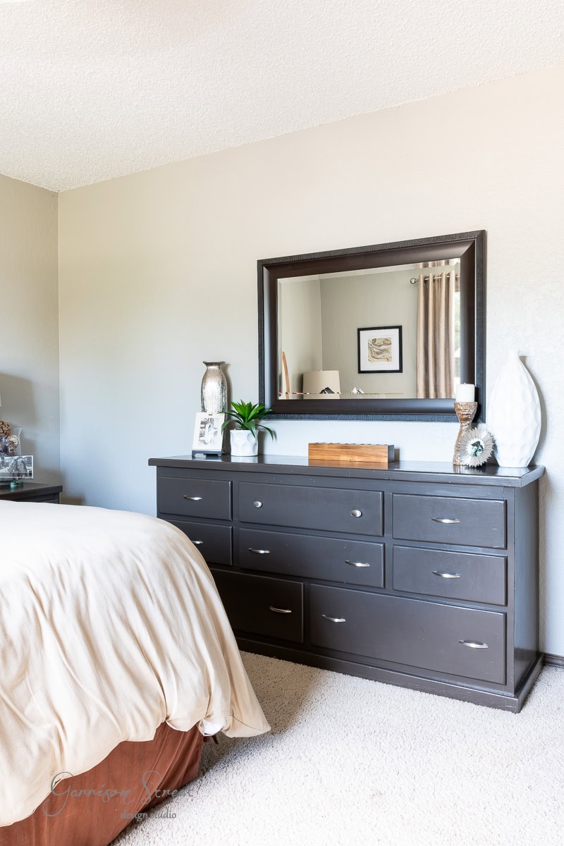 A bedroom features a dark wood dresser paired with a large rectangular mirror. The dresser is adorned with decorative vases, books, and a small plant. Part of the bed with light bedding is visible on the left against neutral-colored walls and carpet.