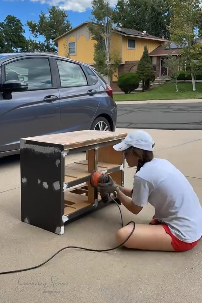 A person in a white cap and red shorts uses a power sander on the wooden surface outside in the driveway. A gray car is parked nearby, while houses and trees provide a serene backdrop.