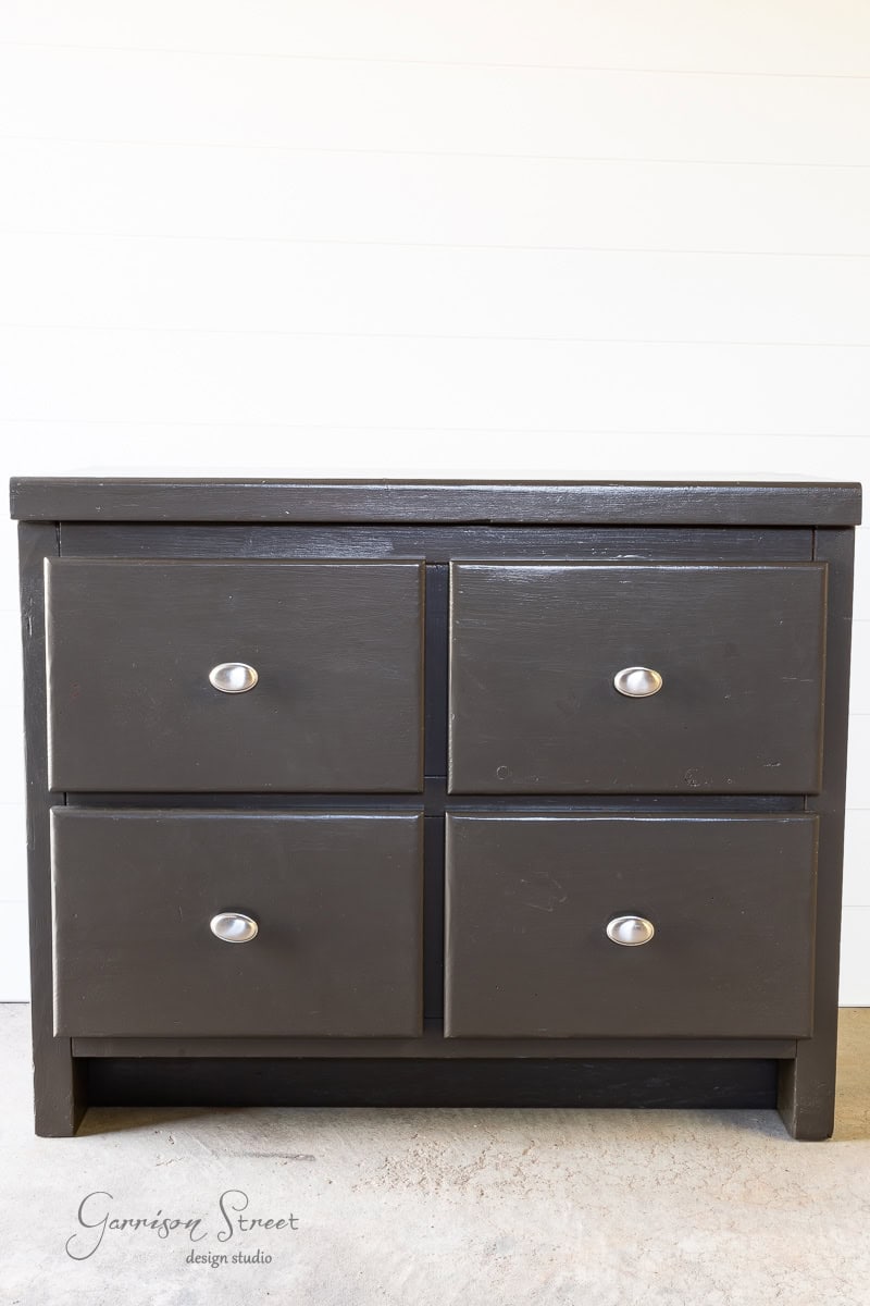 A black wooden dresser with four drawers, each adorned with a round silver knob, stands against a plain white wall on a light-colored floor.