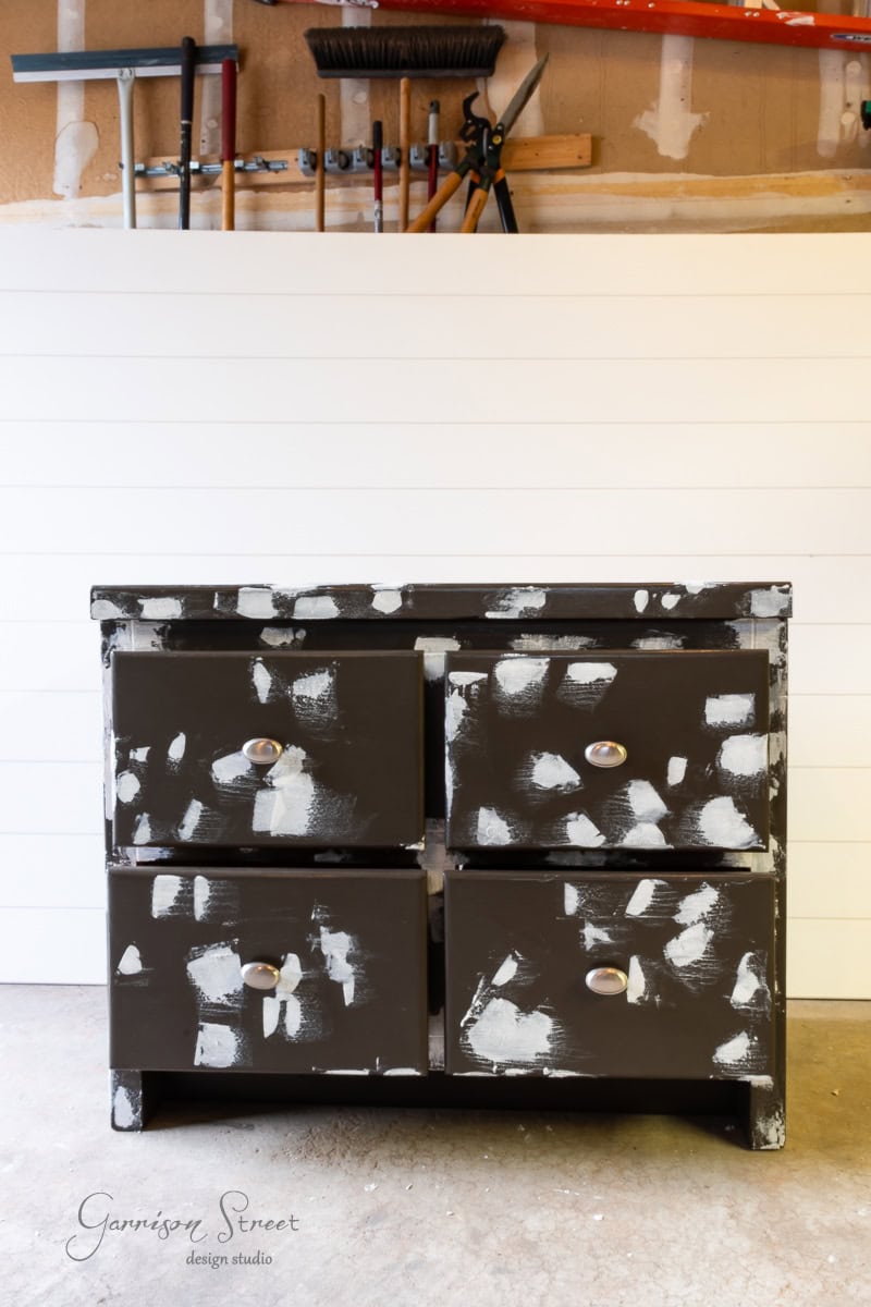 A weathered, dark brown dresser with distressed white paint stands in a garage. It features four drawers with round knobs, and various tools, including a broom and rake, are neatly organized on the wall rack behind it.