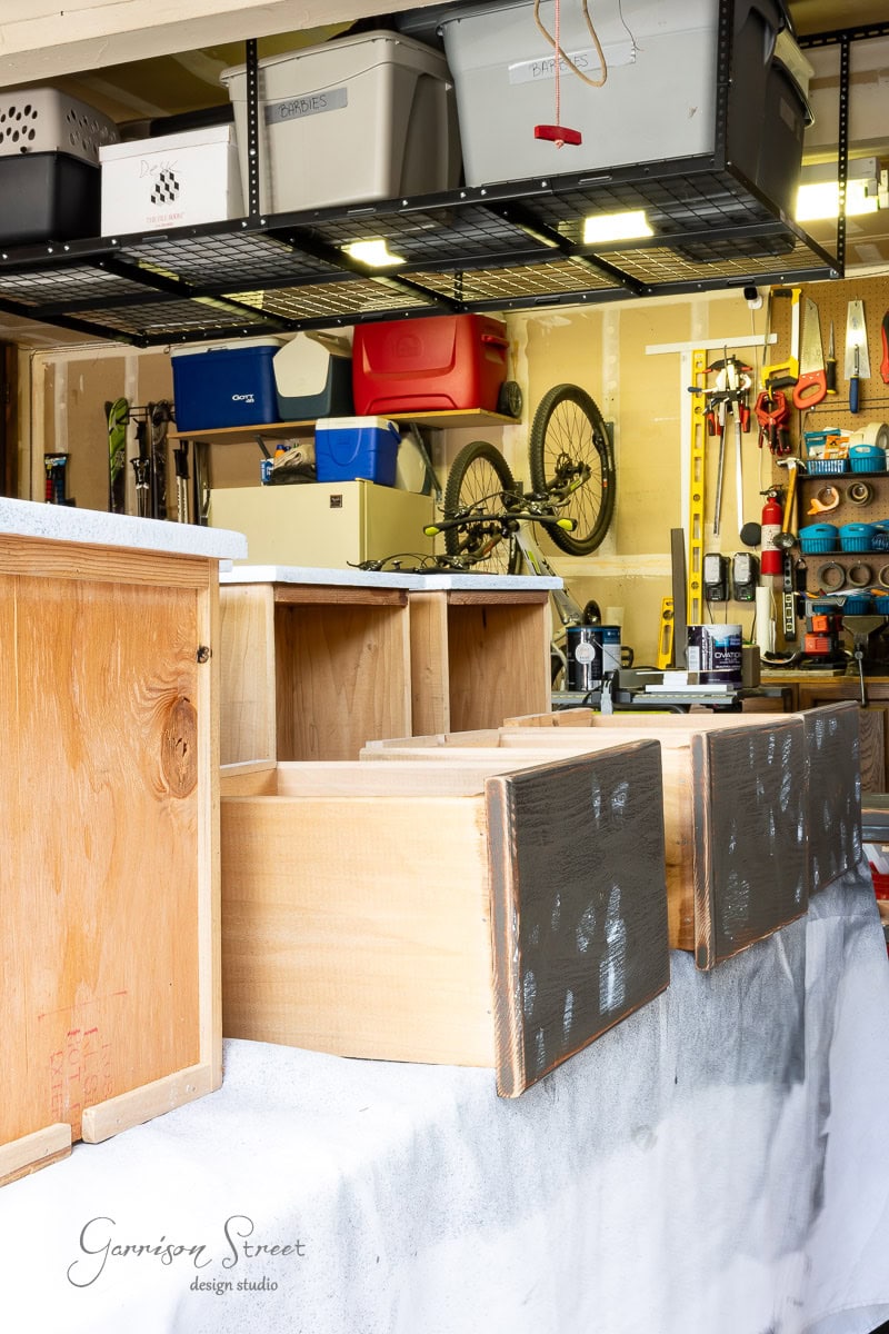 A garage workshop features wooden drawers on a table, partially painted black. Shelves boast plastic storage bins, with tools and a bicycle hanging on the wall.