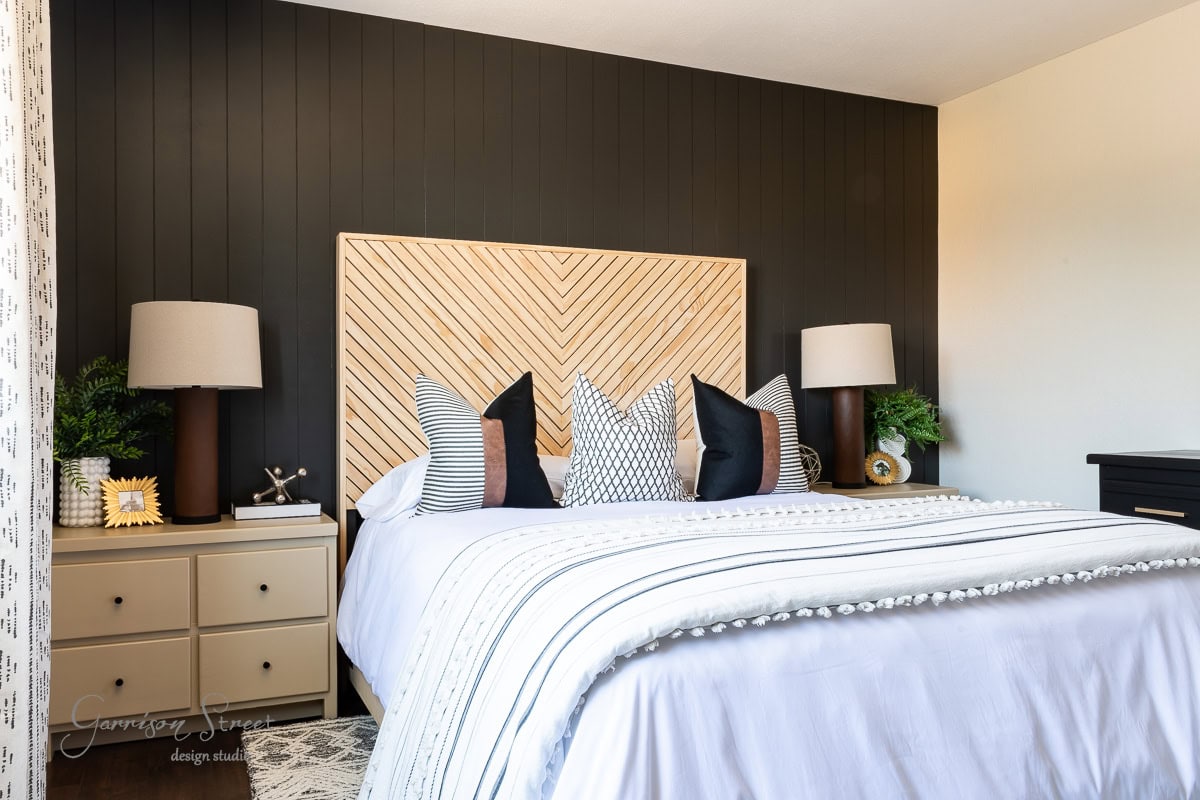 Contemporary guest bedroom makeover featuring a bed with striped bedding and patterned pillows. The headboard boasts a wood chevron design. Two bedside tables each hold a lamp and decor, while a dark accent wall contrasts beautifully with the light room.