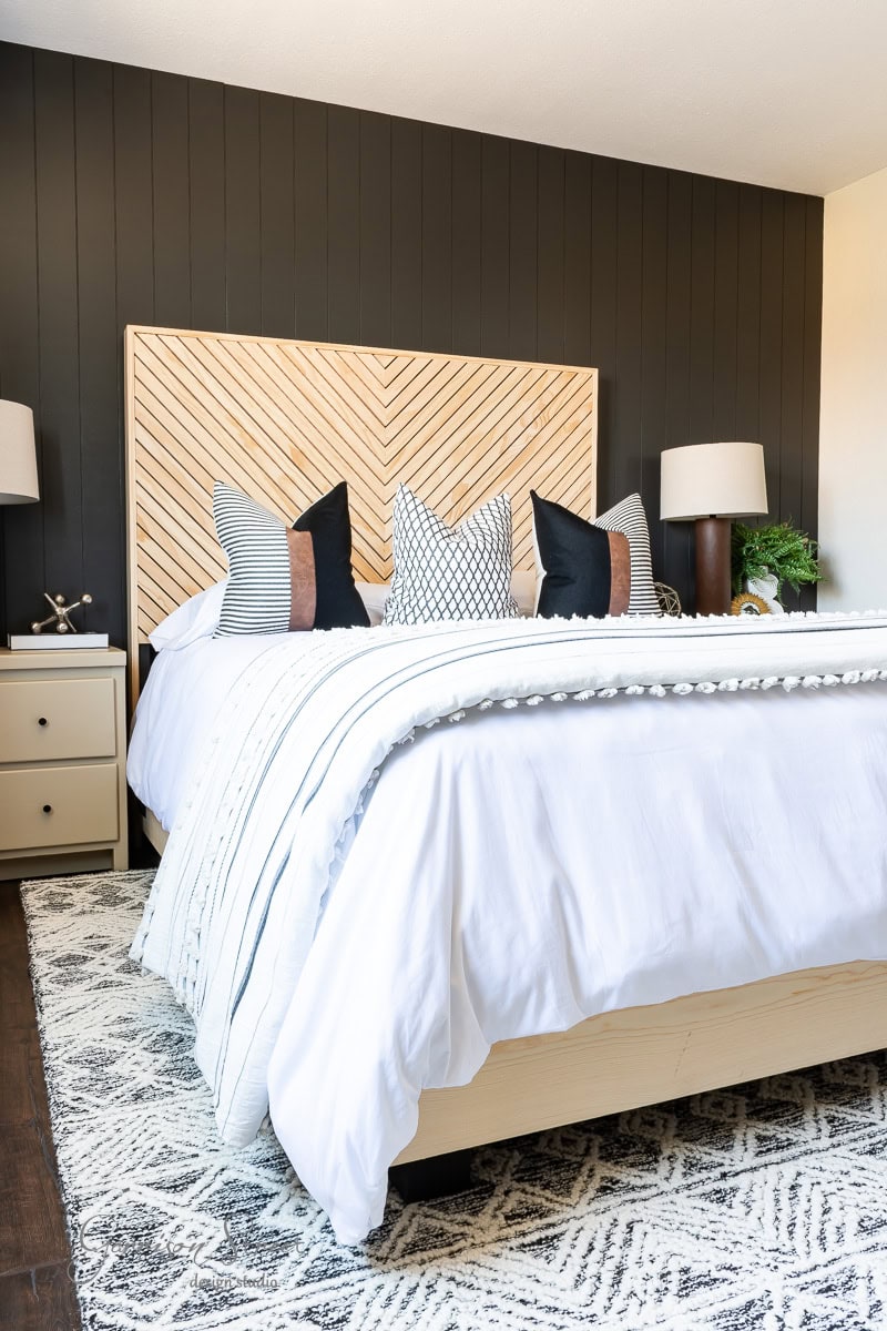 A modern guest bedroom makeover features a large bed with white linens and decorative pillows in various patterns. The headboard boasts a chevron wood design against dark walls. Two nightstands with lamps flank the bed, and a patterned rug completes the look.