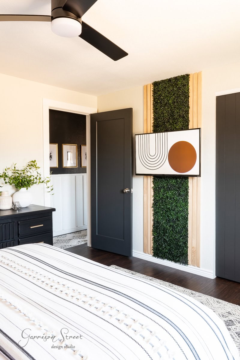 Modern guest bedroom makeover with a geometric wall art piece, black door, and partial green vertical garden panel. The bedspread features striped patterns, and a sleek black ceiling fan is visible. A small dresser holds a plant and decorative items.