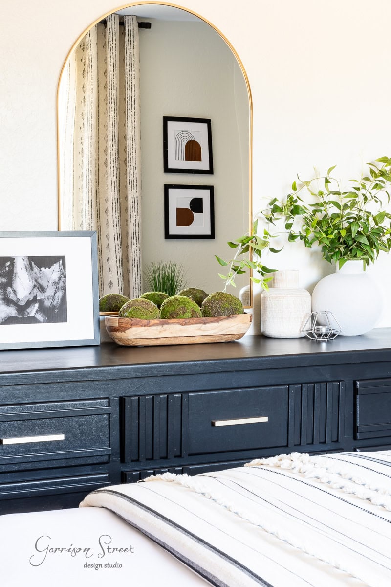 A black dresser topped with a round mirror, decorative framed art, and plants transforms the guest bedroom makeover. A wooden bowl with moss balls adds greenery, while the reflection reveals curtains and more wall art. A striped blanket is partially visible in the foreground.