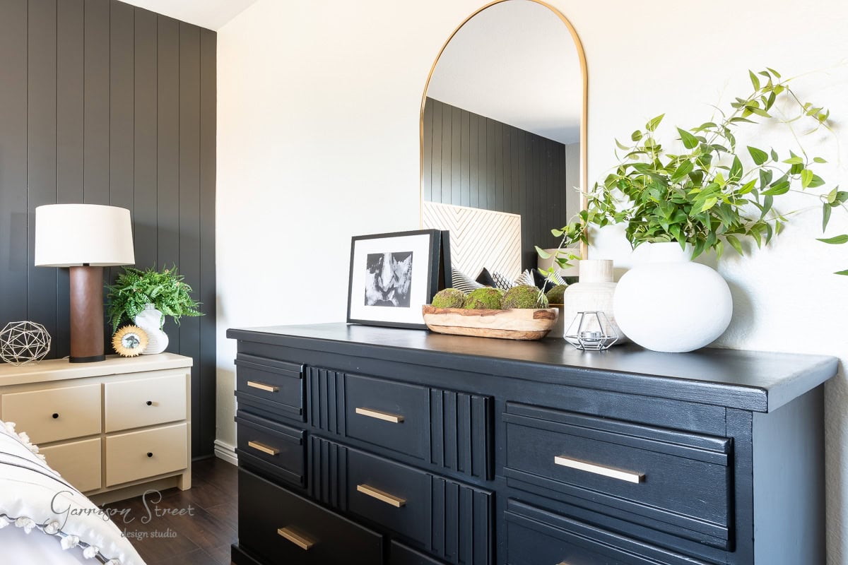 Experience a guest bedroom makeover with a modern setup featuring a dark dresser with sleek handles. Decorative items include a round mirror, a framed photo, and vases with green plants. In the background, find a beige nightstand adorned with a lamp and decorative sphere.