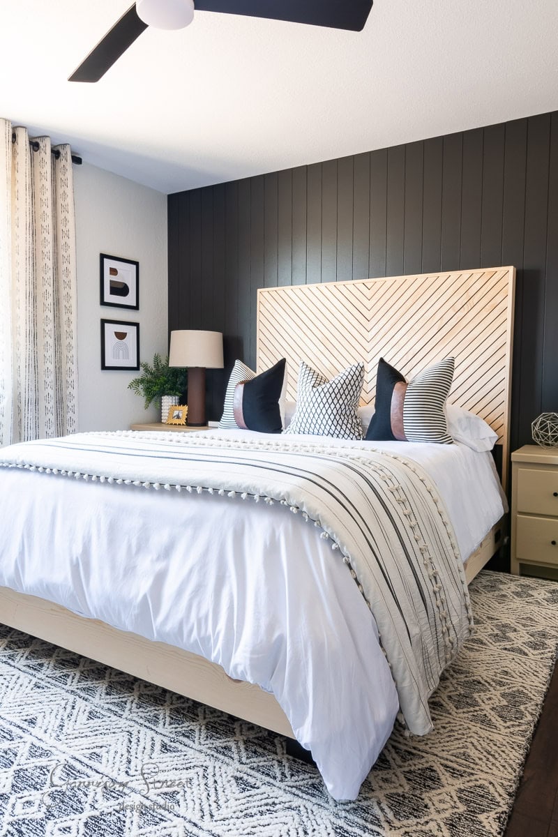 A modern guest bedroom makeover showcases a large bed with a patterned headboard and striped pillows. The space features dark accent walls, light curtains, and framed pictures. A geometric rug adorns the floor while a ceiling fan adds function and style above.