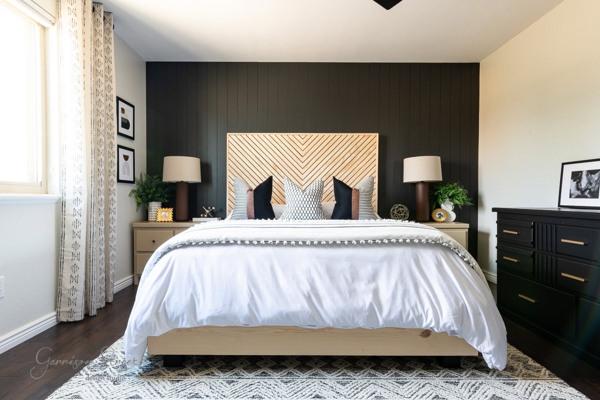 A modern guest bedroom makeover showcases a large bed with a white comforter and decorative pillows against a dark accent wall. Flanked by matching nightstands with lamps and plants, the room includes a window with patterned curtains and a dresser adorned with framed photos.