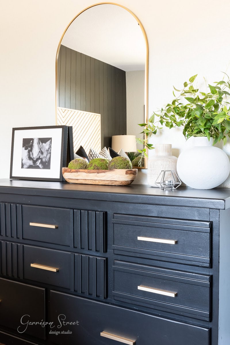 A stylish black dresser with silver handles, perfect for a guest bedroom makeover, is topped with a leafy plant in a gray vase, decorative items, and framed art. A large arched mirror reflects part of the room, adding depth to the modern decor.