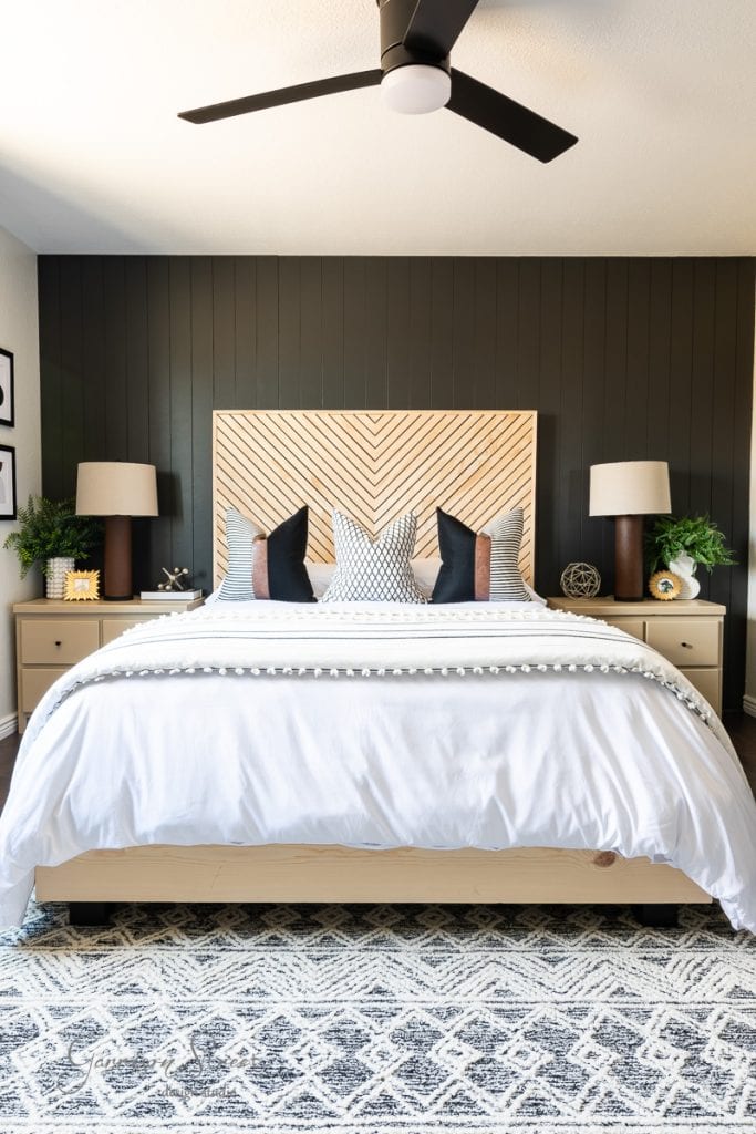 A modern guest bedroom makeover features a white bed, black and white accent pillows, and a chevron wood headboard against a dark paneled wall. Two wooden nightstands with lamps and plants flank the bed, while a patterned rug covers the floor beneath the ceiling fan overhead.