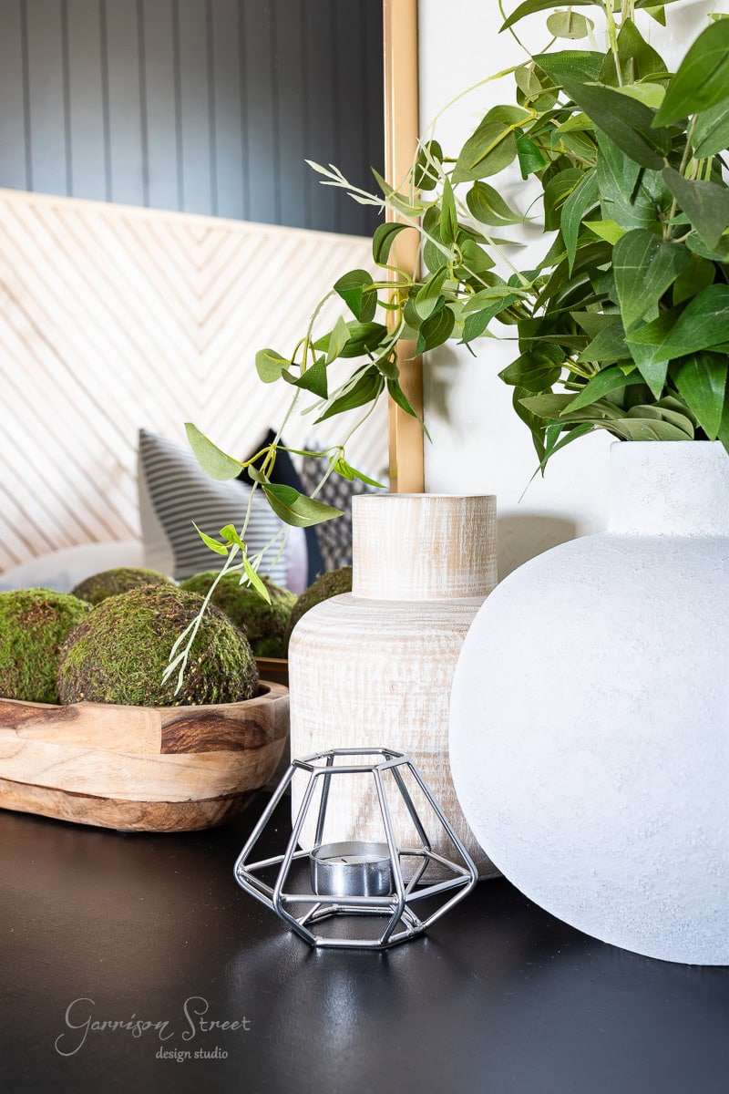 A stylish guest bedroom makeover scene features two vases and a candle holder on a dark surface. One vase is textured off-white, and the other is wood-toned, decorated with leafy green branches. A wooden bowl holds green moss balls on the left.