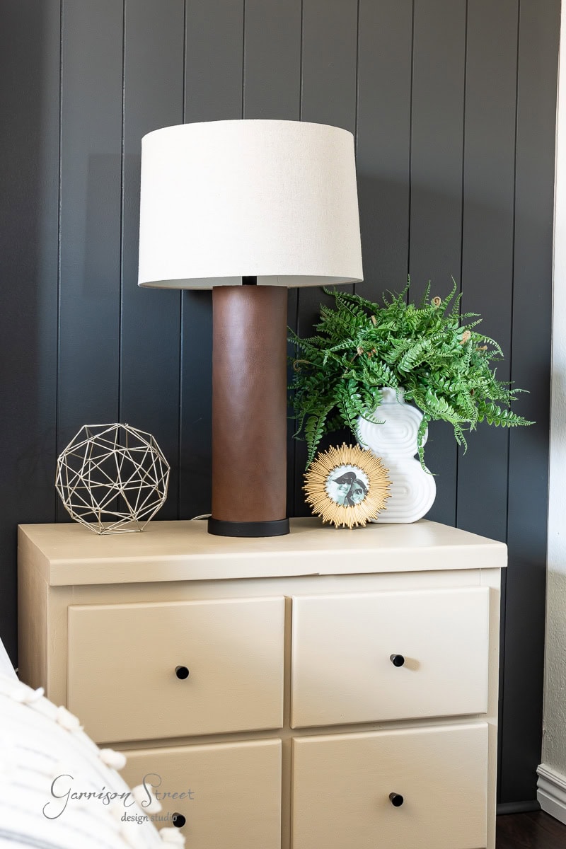 A modern bedside table enhances the guest bedroom makeover, featuring a beige lamp, geometric decor, a green plant in a white vase, and a small framed photo. The table's two drawers have black knobs against the backdrop of a dark paneled wall.