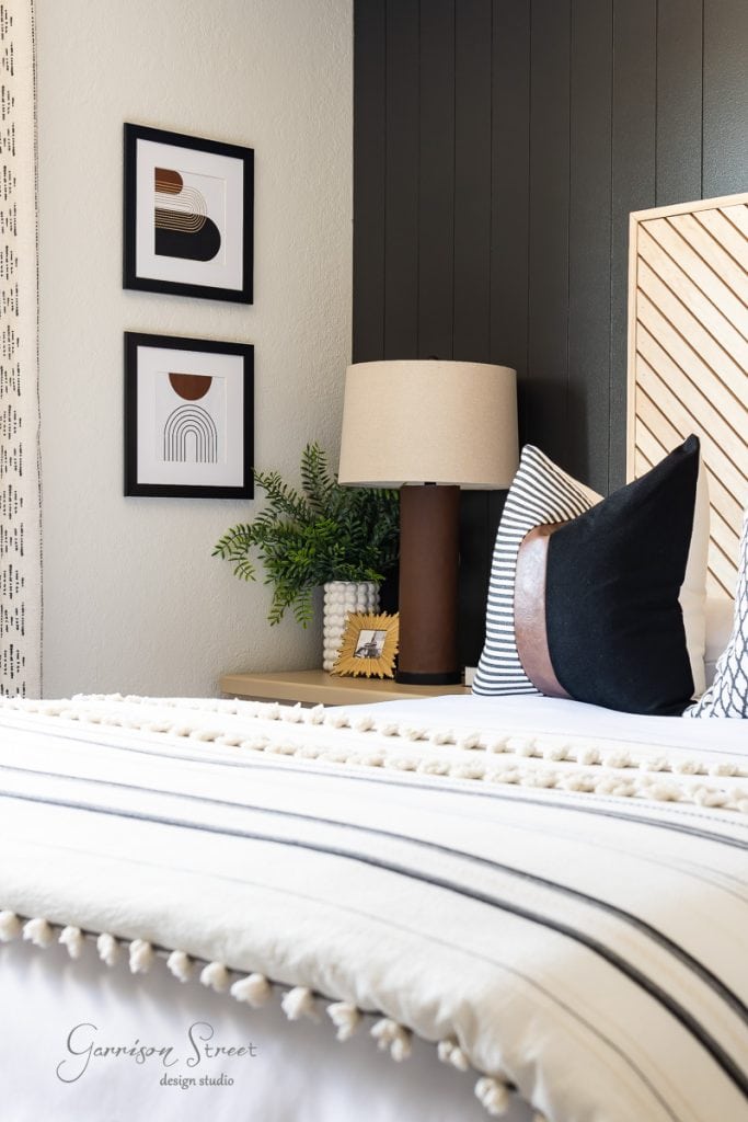 A cozy guest bedroom corner features a bed with white and black striped bedding and pom-pom details. A wooden nightstand holds a lamp and plant, while two black-and-white abstract artworks adorn the wall above, complemented by a dark accent panel.