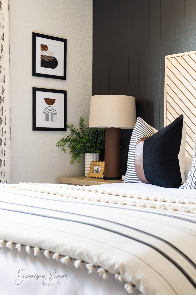 A cozy guest bedroom corner features a bed with white and black striped bedding and pom-pom details. A wooden nightstand holds a lamp and plant, while two black-and-white abstract artworks adorn the wall above, complemented by a dark accent panel.