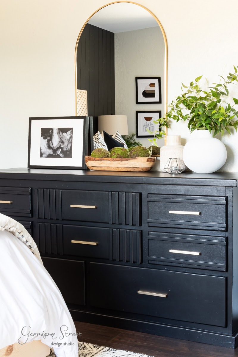 A black dresser with six drawers anchors the guest bedroom makeover, topped with decorative items like a mirror, framed artwork, and a plant. A wooden tray holds moss and a small lantern, while the wall behind showcases additional framed art for a cozy, inviting atmosphere.