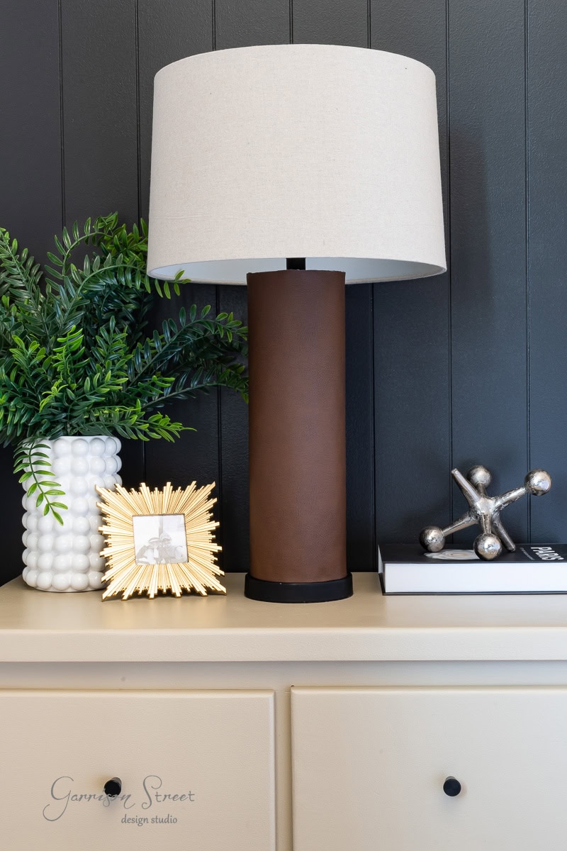 A modern sideboard perfect for a guest bedroom makeover, featuring a brown cylindrical lamp with a beige shade, a white textured vase filled with green foliage, a small gold picture frame, and a silver decorative sculpture of interconnected spheres. The backdrop is vertical dark paneling.