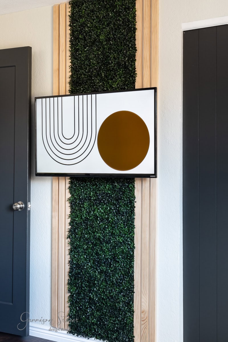 A modern art piece with black lines and a brown circle is mounted on a wall, adding a contemporary touch to the guest bedroom makeover. The artwork stands out against vertically arranged wooden panels and artificial greenery, while two dark doors elegantly flank the installation.