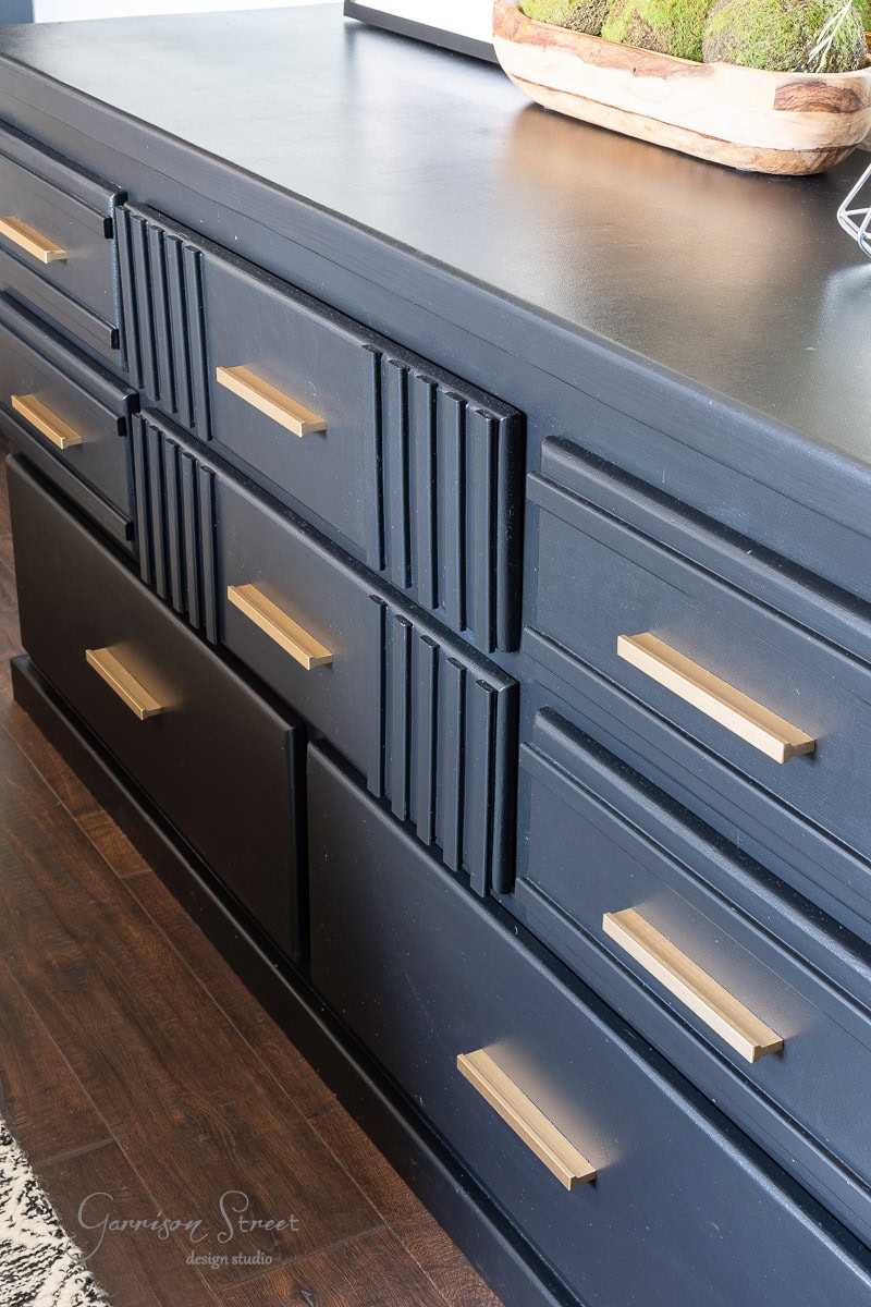 A stylish black dresser with gold handles and a textured design on the upper drawers adds elegance to your guest bedroom makeover. The dresser stands on a dark wood floor and is topped with decorative items, including a planter with moss and a geometric ornament.