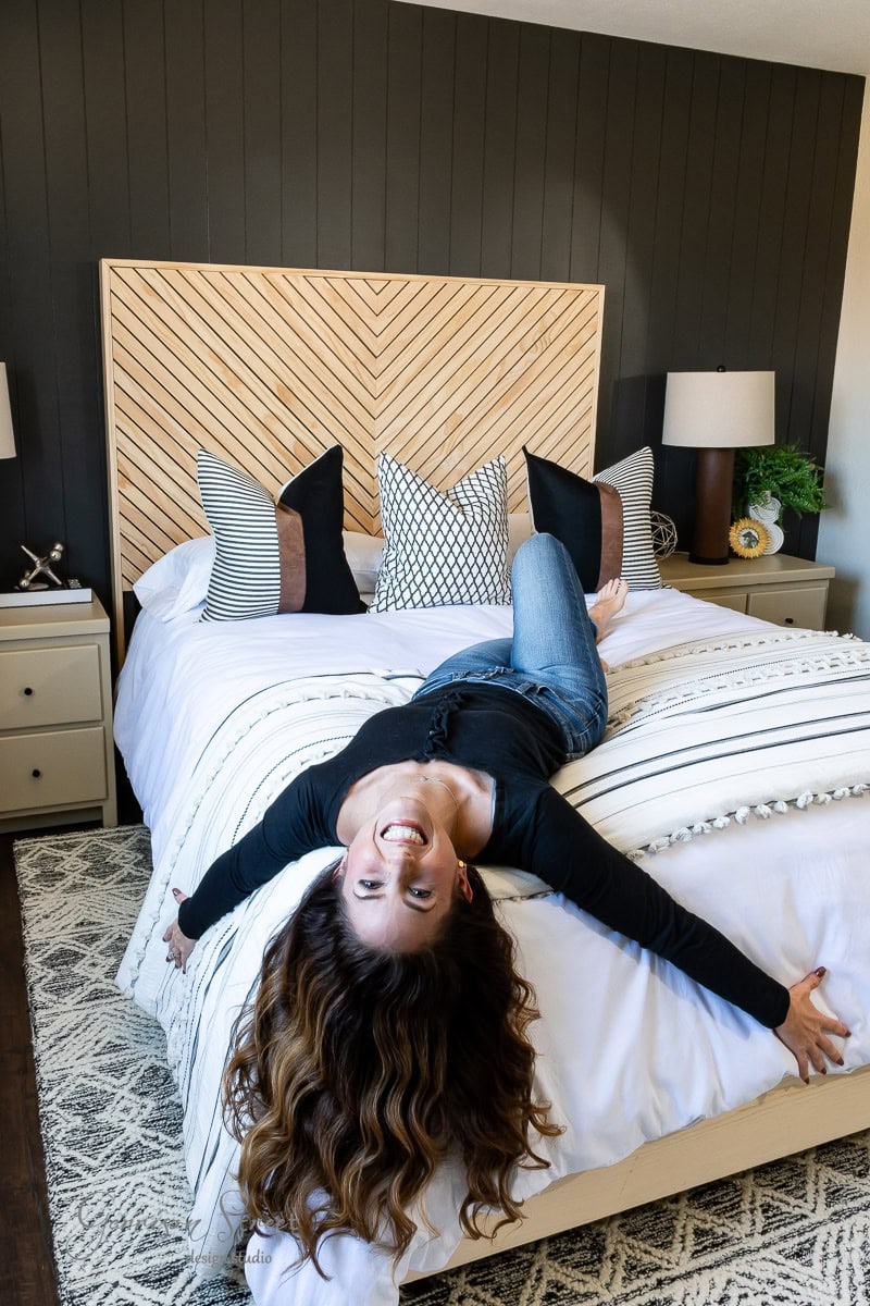 A woman with long, wavy hair is playfully lying upside down on a bed in the newly transformed guest bedroom. The room boasts a patterned headboard, multiple throw pillows, bedside tables with lamps, and a textured rug. She's smiling and wearing a casual outfit.
