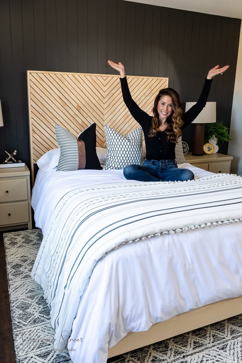 A woman sits joyfully on a bed with arms raised, reveling in the guest bedroom makeover. The bed is adorned with decorative pillows and a striped comforter. A chevron-patterned headboard adds flair, while two lamps perch on nightstands beside the bed.
