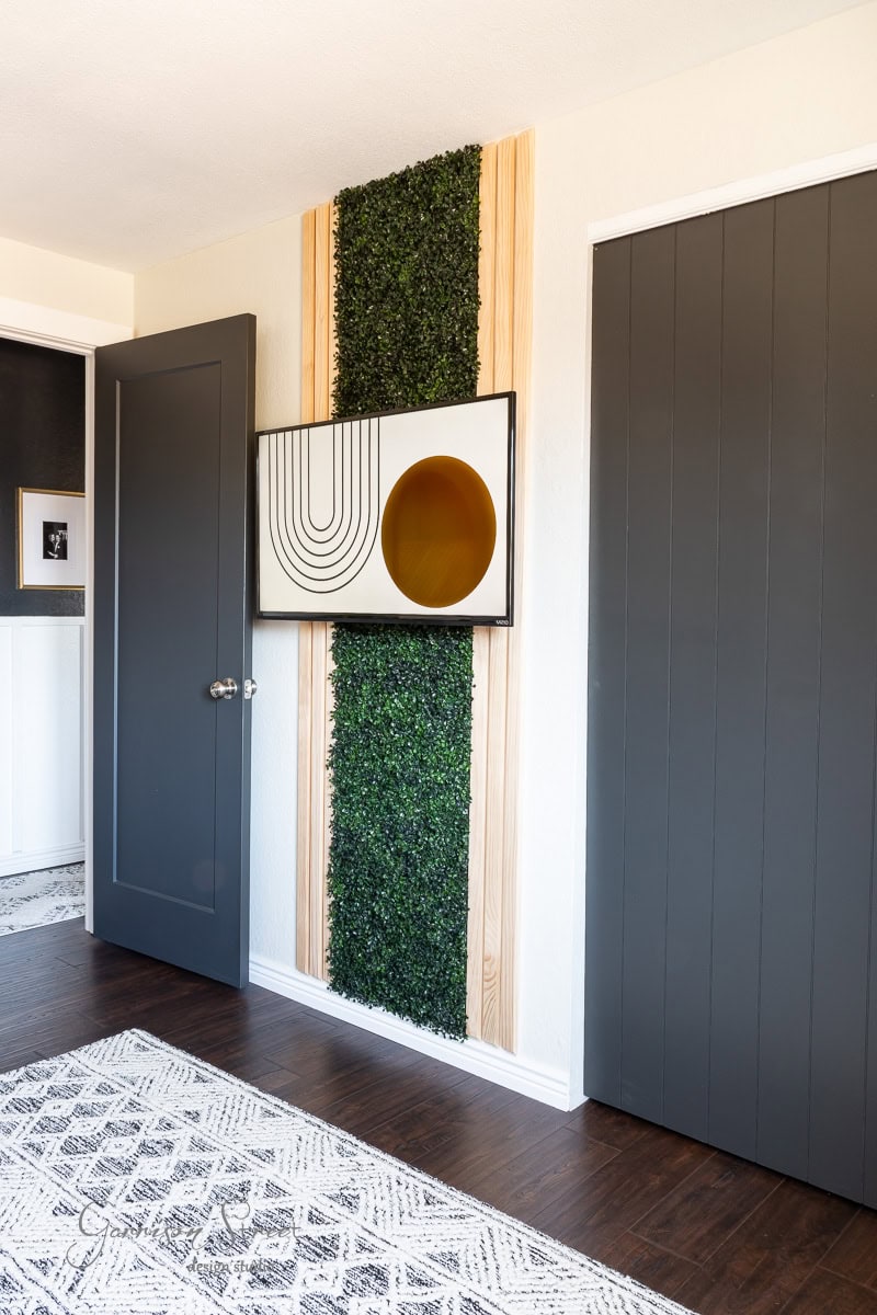 A modern guest bedroom makeover showcases a vertical green plant wall with wood panels, a framed abstract artwork, dark gray doors, and a geometric patterned rug on dark wood flooring.