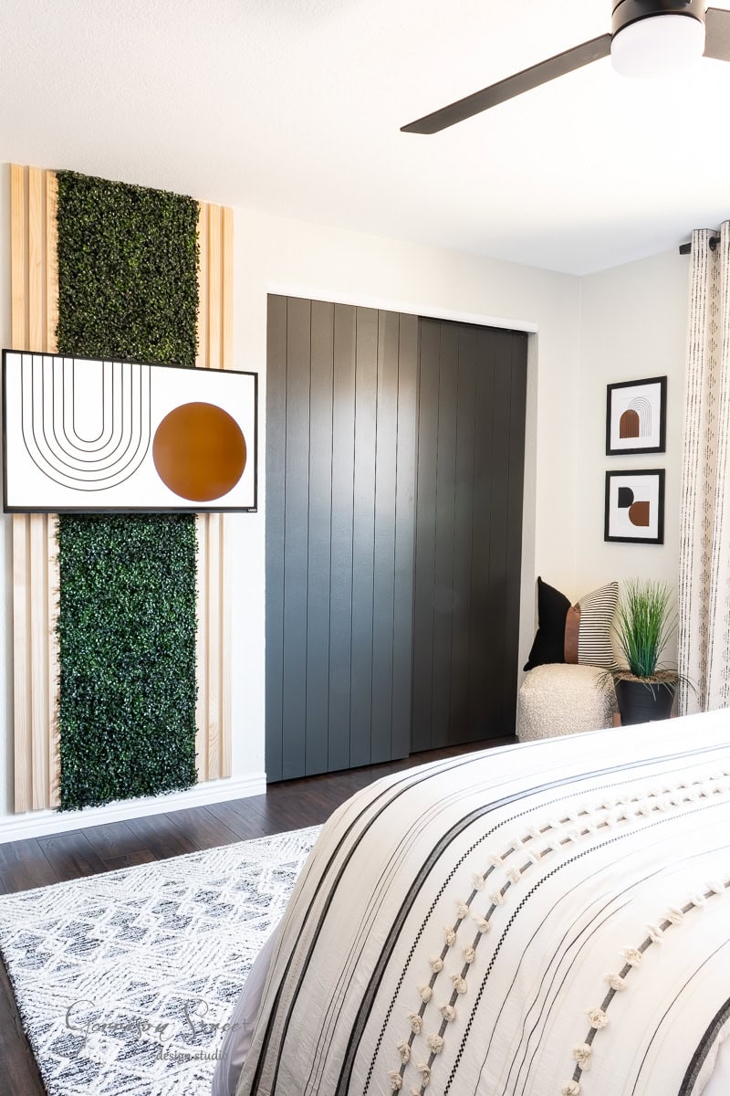 A modern guest bedroom makeover features a large bed with striped linens, a wall-mounted TV, and a vertical plant panel. A dark sliding door and abstract framed art enhance the contemporary design. A small chair with cushions sits in the corner, near a window.