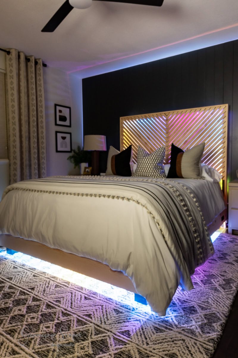 A modern guest bedroom makeover showcases a large bed with decorative pillows and a textured duvet. The bed frame is illuminated by colorful LED lights, while dark accent walls, framed art, and patterned curtains complement a geometric rug on the floor.