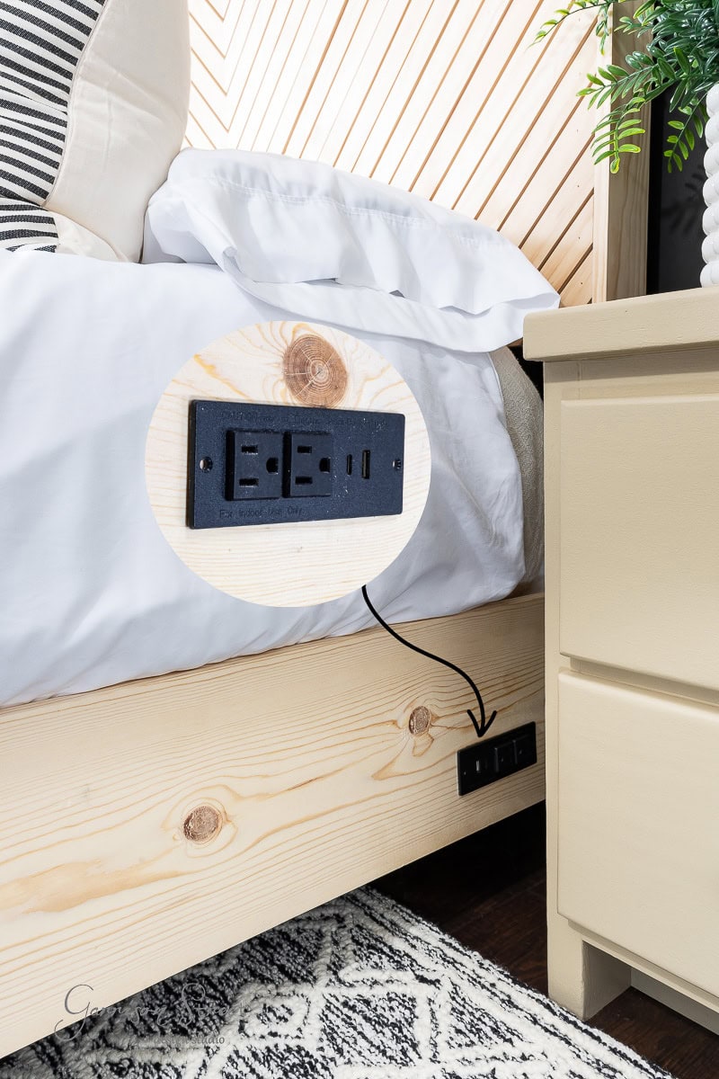 A wooden bed frame with built-in electrical outlets, featuring two sockets and USB ports, is perfect for your guest bedroom makeover. The outlets are highlighted with a circular inset. Nearby, a beige nightstand complements the sleek design alongside a portion of a white and black rug.