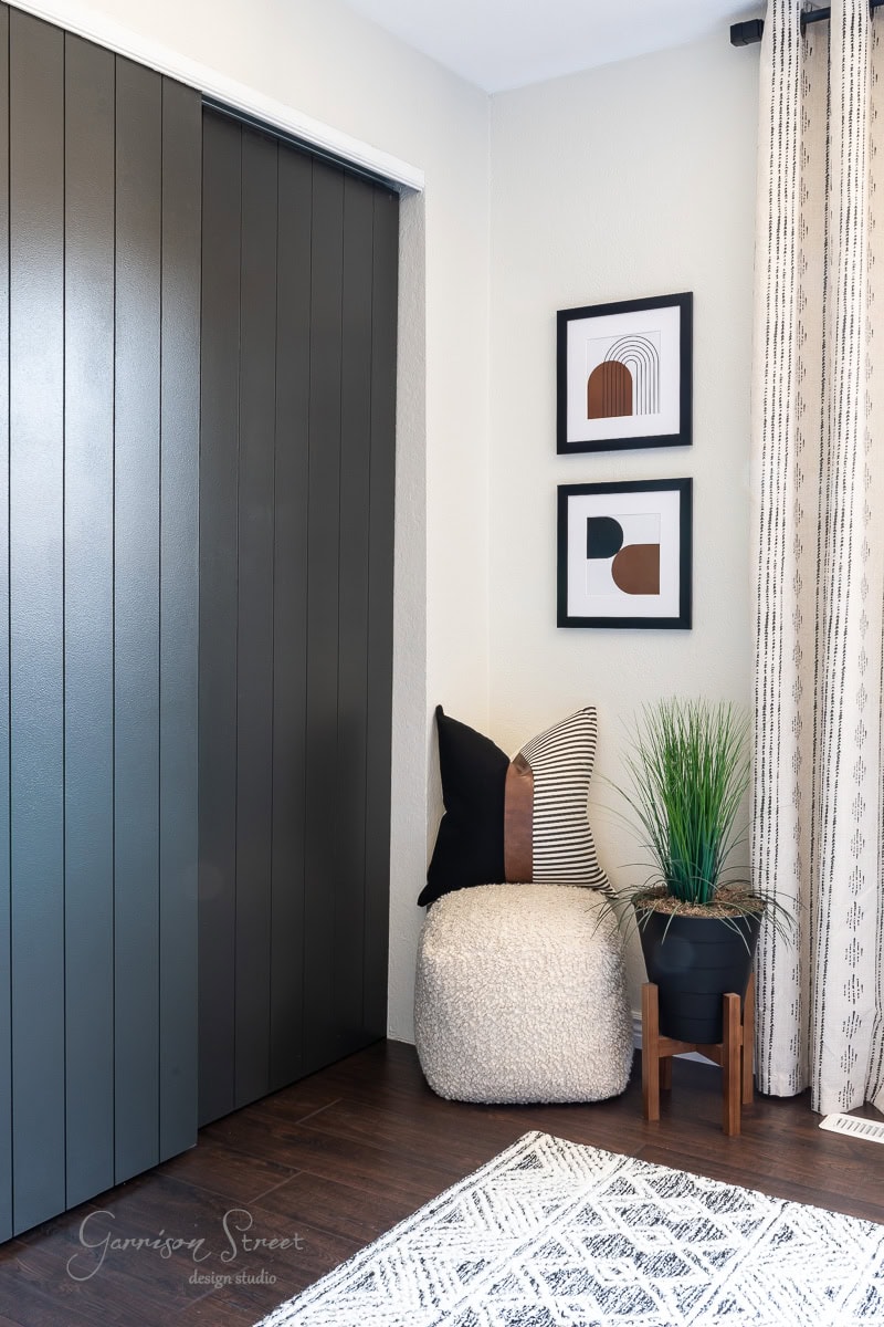 A modern guest bedroom makeover features black paneled sliding doors, a beige pouf with striped pillows, a plant in a black pot on a wooden stand, and two geometric art pieces on the wall. Cream curtains and a patterned rug complete this cozy corner.