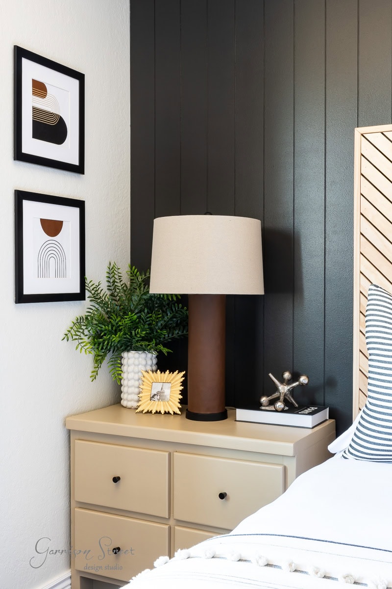 A modern guest bedroom makeover features a beige nightstand with a table lamp, decorative objects, and framed abstract art on the wall. The dark-painted wall contrasts with light bedding and furniture, while a potted plant adds a refreshing touch of greenery.
