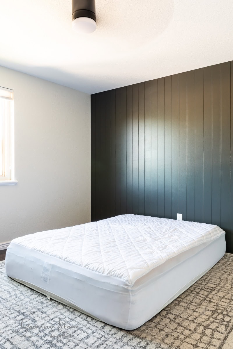 A minimalist bedroom highlights a DIY full-size bed frame with a mattress covered by a white quilted pad on a gray-patterned rug. The room showcases a dark vertical paneled accent wall, with light streaming through the window on the left and a ceiling light fixture centered above.