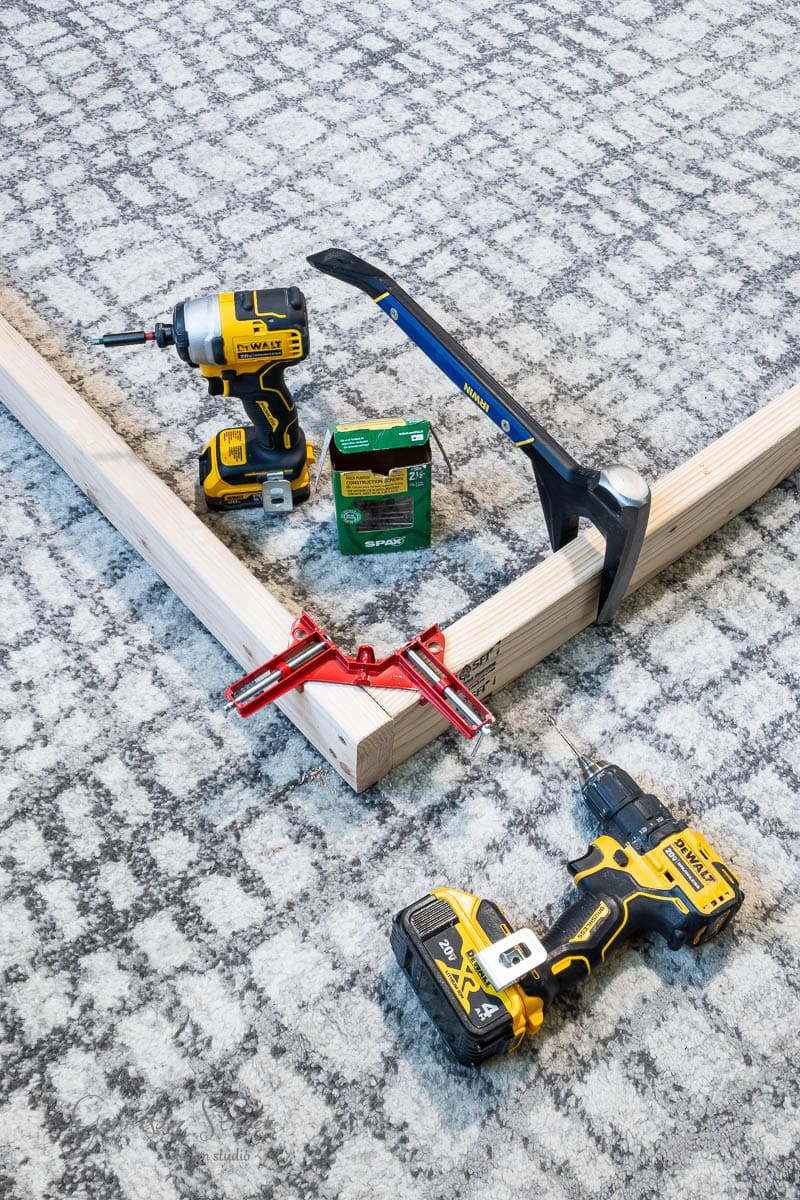 Assorted power tools and accessories on a gray patterned carpet: two yellow cordless drills, a blue pry bar, a green box of screws for your DIY full-size bed frame project, a red angle clamp, and wooden beams forming a right angle.