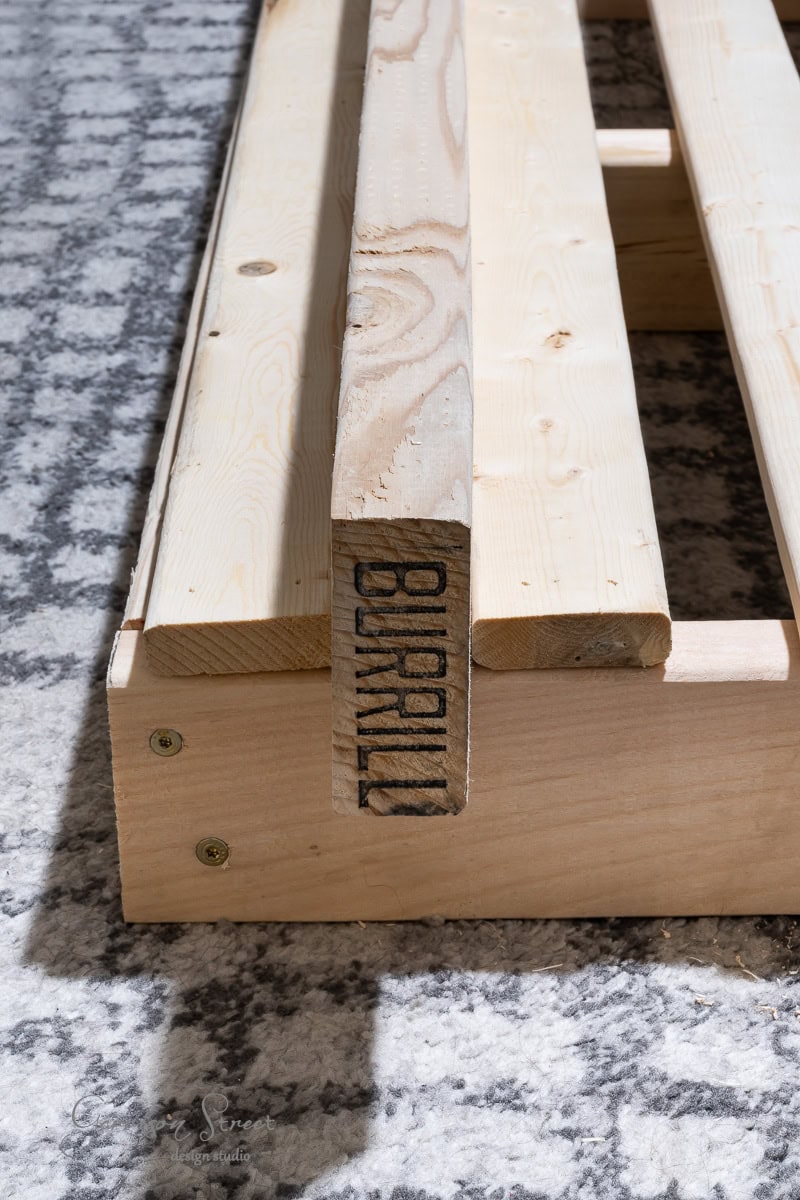 A close-up of a wooden pallet on a carpeted floor, ideal for a DIY full-size bed frame. The pallet is made of light-colored wood with the word "SLURMIL" stamped on one of the slats. Two metal screws are visible on the side.