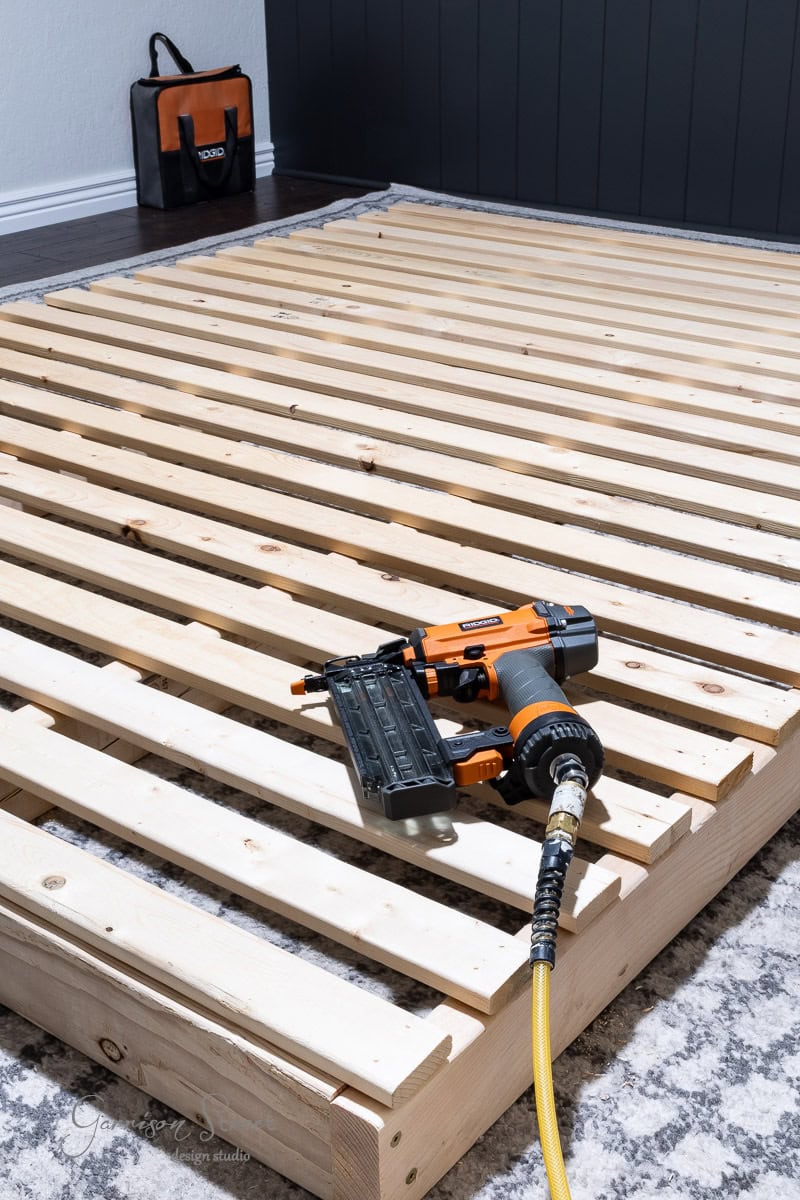 A DIY full-size bed frame takes shape with evenly spaced slats on a wooden platform. An orange and black nail gun rests on the frame, connected to an air hose. In the background, a matching black and orange tool bag sits on the carpeted floor, ready for action.