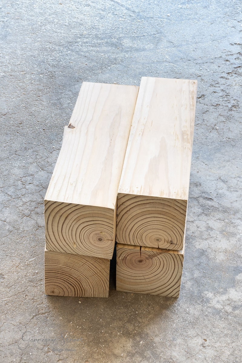 Four stacked, freshly cut wooden beams with visible tree rings are arranged on a textured concrete surface, perfect for crafting a DIY full-size bed frame.