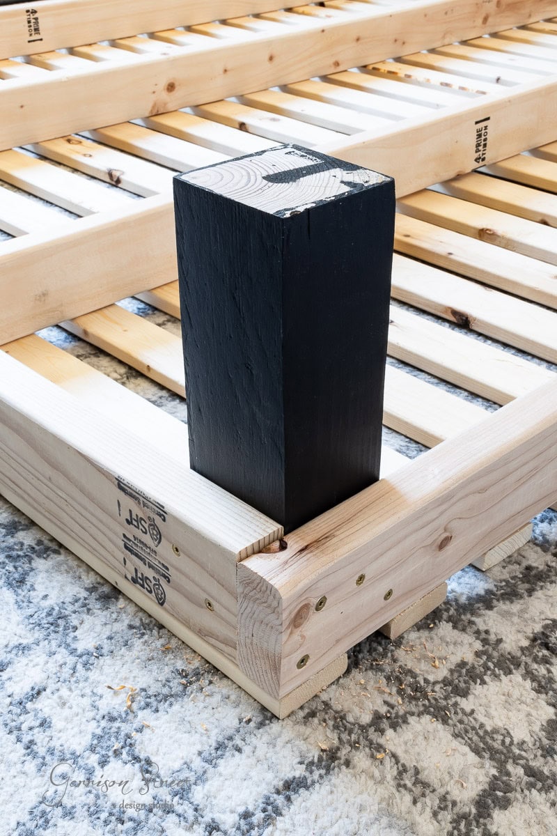 DIY enthusiasts will appreciate the wooden bed frame on a patterned rug, featuring sturdy slats and a large black wooden leg attached at the corner with metal brackets and screws. Perfect for those looking to craft their own full-size centerpiece in any bedroom.
