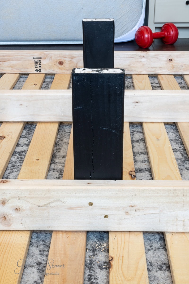 Wooden slats arranged parallel on a textured floor form the base of a DIY full size bed frame, with a black rectangular block in the center. A red dumbbell is visible in the background, partially obscured by a white mattress.