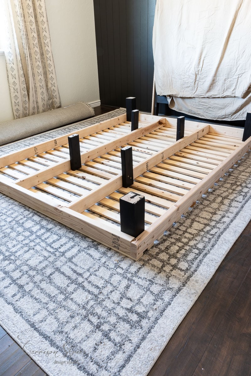 A partially assembled DIY full size bed frame lies on a gray and white patterned rug in the room. Featuring multiple wooden slats and black support posts, it sits near a rolled-up rug and draped curtains visible in the background.