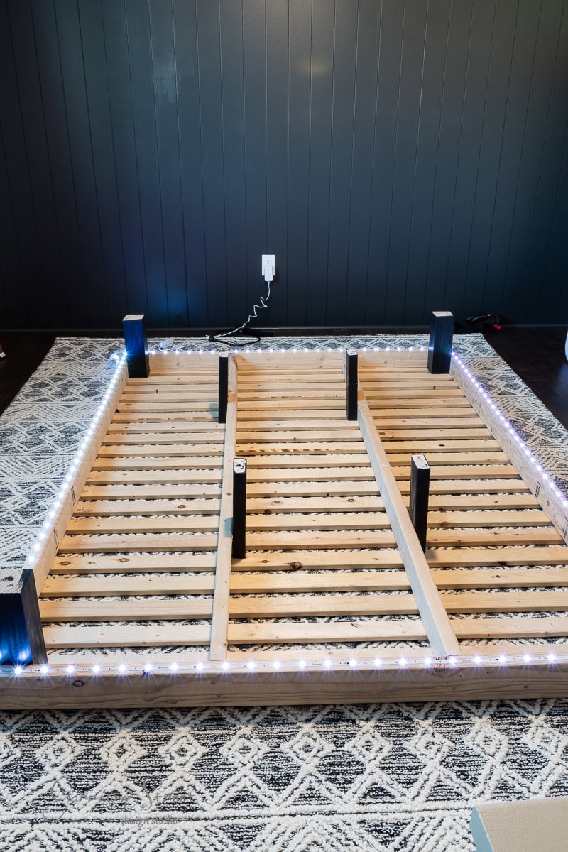 A DIY full-size bed frame with slats is lying upside down on a patterned rug. LED strip lights adorn its perimeter, casting a glow. A black padded headboard stands against the dark wall in the background, while several bed legs are evenly spaced out.