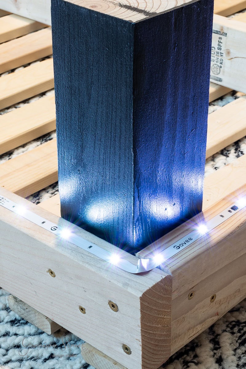 A DIY full size bed frame with a dark vertical post is illuminated by LED strip lights attached along its edge. Resting on a patterned rug, the cool white glow of the lights highlights the structure's corner, adding a modern touch to this handmade creation.
