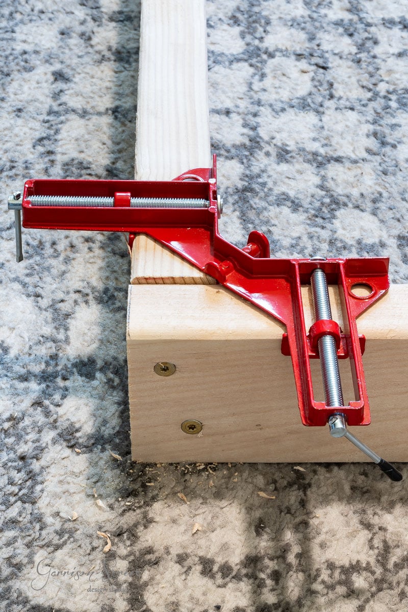 A red woodworking corner clamp secures two wooden boards at a right angle on a textured gray surface, an essential tool for any DIY full-size bed frame project. The clamp features silver screws, while the boards are fixed with gold screws.