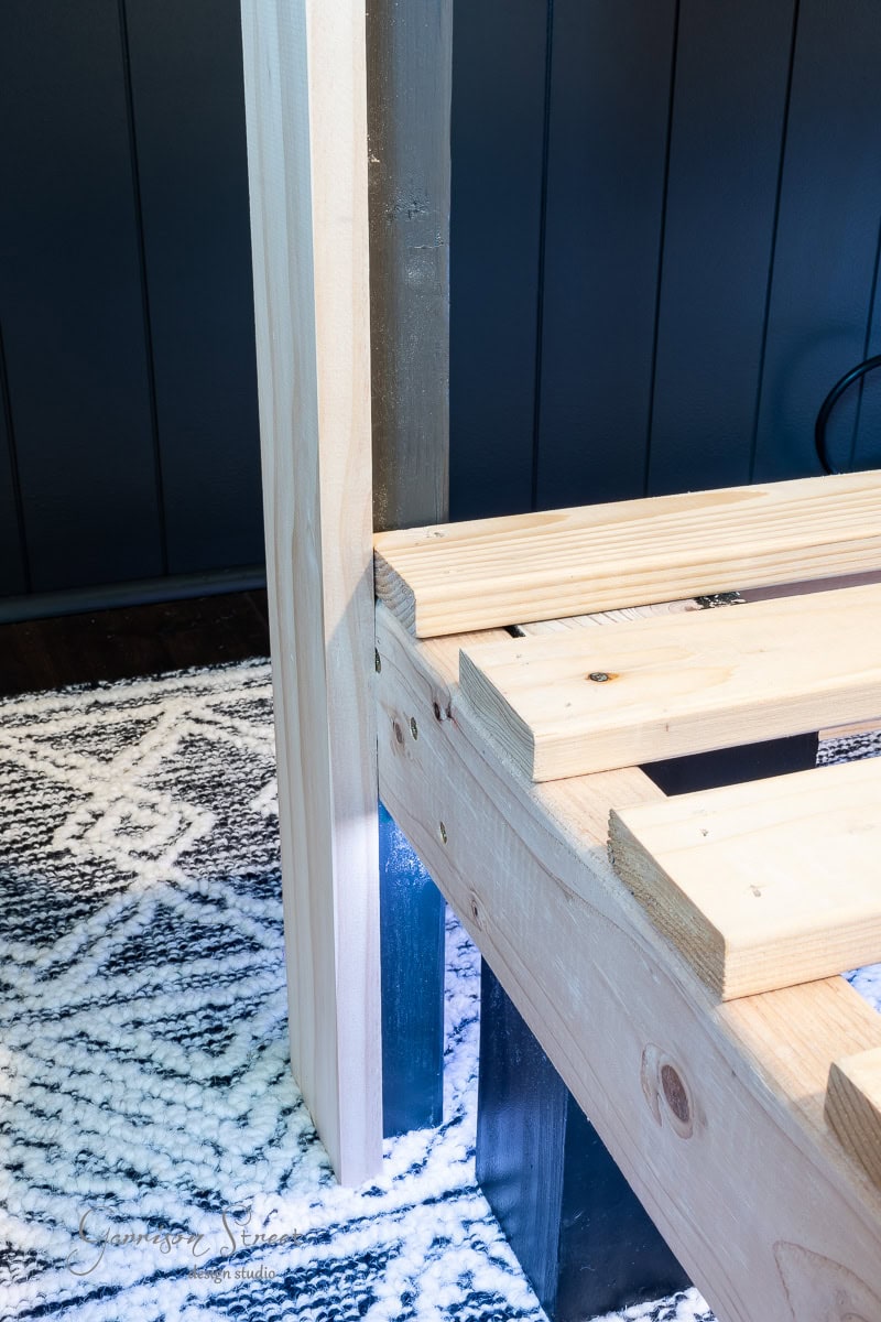 A DIY full size wooden bed frame in progress rests on a patterned rug. It features vertical and horizontal planks connected with screws, offering a stunning contrast between the light wood and dark wall in the background.